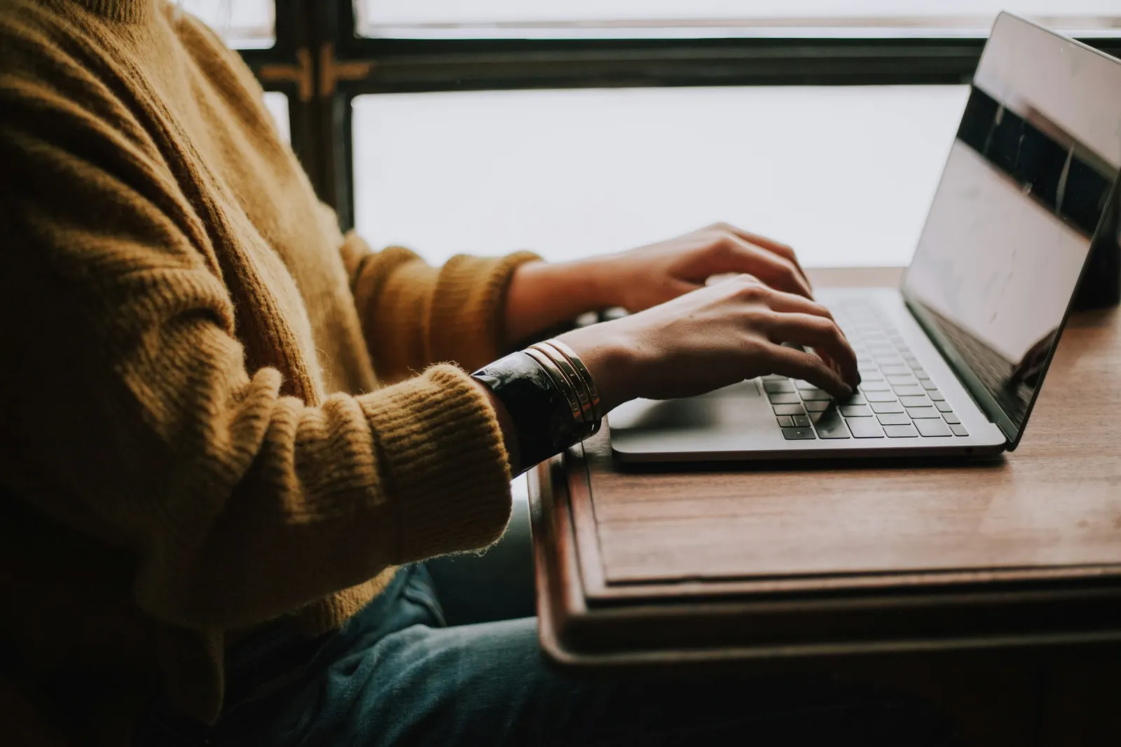 Hände tippen auf einem Laptop an einem Holztisch am Fenster